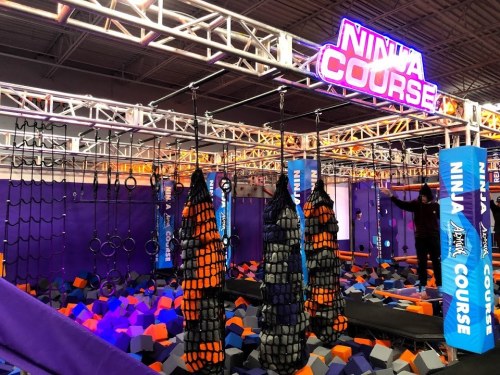 An indoor ninja course featuring climbing nets, rings, and a foam pit, with vibrant purple and orange colors.