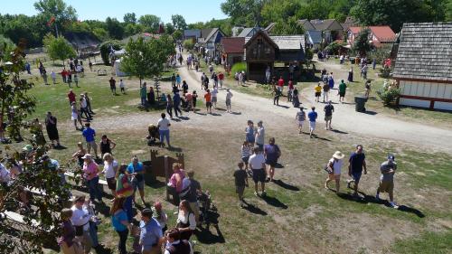Aerial view of a bustling outdoor festival with people walking along paths and various buildings in a green landscape.
