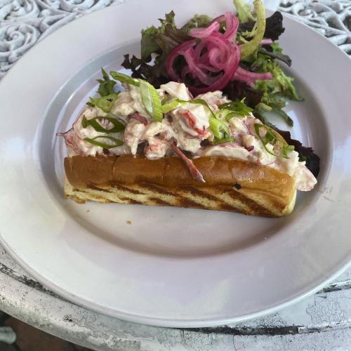 A lobster roll topped with lettuce and a side of mixed greens and pickled onions on a white plate.