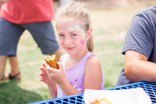 A young girl with face paint smiles while holding a corn dog at a picnic table outdoors.
