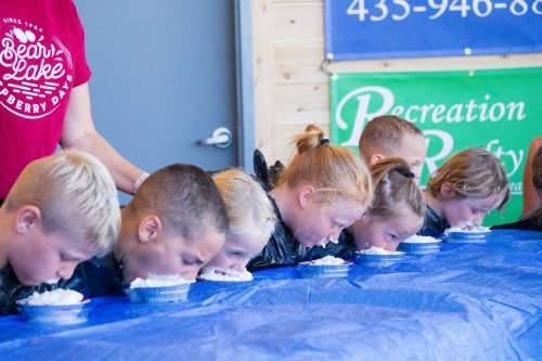 Children compete in a pie-eating contest, leaning over a table covered with blue cloth, with whipped cream pies in front of them.