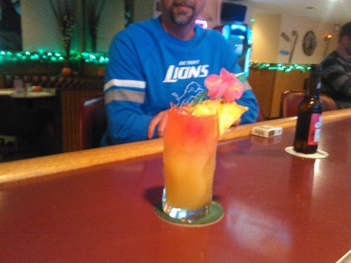 A colorful cocktail with a pineapple slice and flower sits on a bar, with a man in a blue shirt in the background.