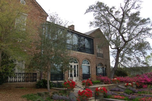 A brick house with large windows, surrounded by colorful flowers and trees, set against a cloudy sky.