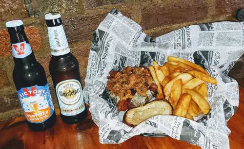 A basket of fries and a sandwich topped with crispy onions, accompanied by two bottles of beer on a wooden table.
