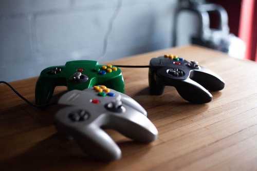 Three video game controllers in green, silver, and black resting on a wooden surface.