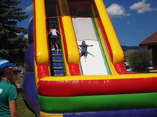 Two children play on a colorful inflatable slide on a sunny day, with one sliding down and the other climbing up.