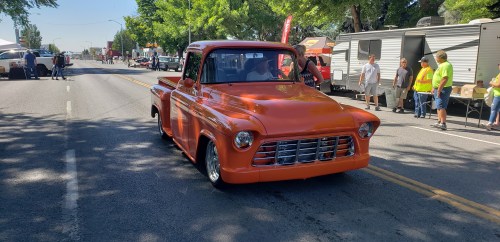 An orange vintage pickup truck drives down a tree-lined street during a car event, with people and booths in the background.