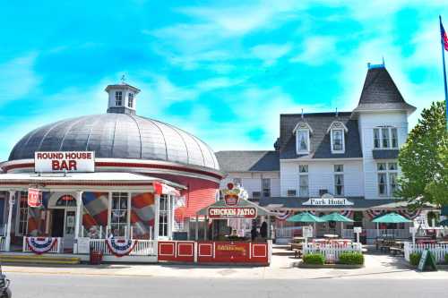 A colorful round building labeled "Round House Bar" next to a large Victorian-style hotel with outdoor seating.