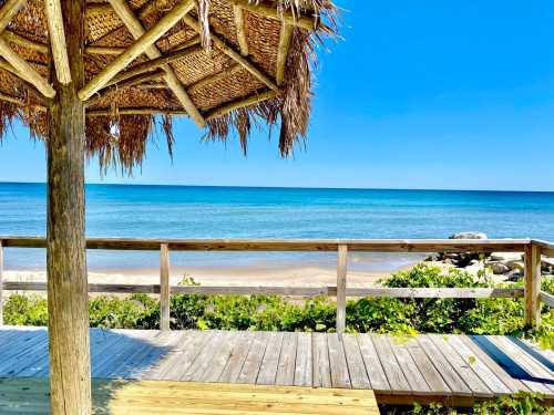 A beach view from a wooden deck with a thatched umbrella, overlooking calm blue waters and a sandy shore.