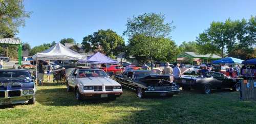A car show featuring classic cars, tents, and attendees in a grassy park setting on a sunny day.