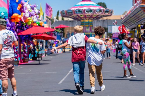 Two friends walk arm in arm down a vibrant fairway filled with colorful attractions and people enjoying the festivities.