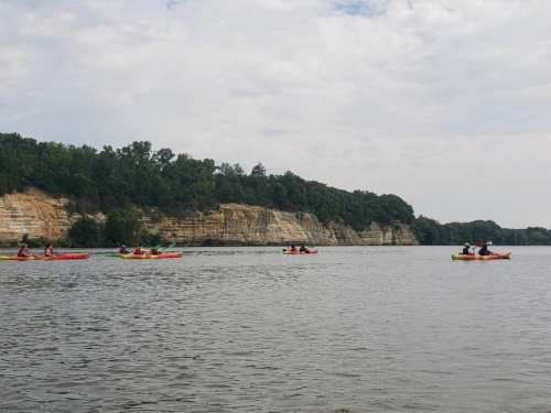 A group of kayakers paddles along a calm river with rocky cliffs and trees in the background under a cloudy sky.