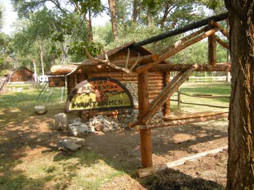 Log cabin structure with a sign reading "Fort Buenaventura" and "Mount Men" in a grassy area surrounded by trees.