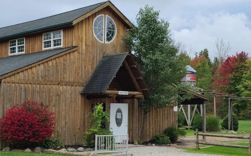 A rustic wooden building with a round window, surrounded by greenery and colorful autumn trees.