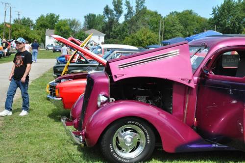 A classic car show featuring a purple vintage car with its hood open, surrounded by other classic vehicles and spectators.