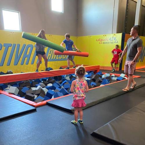 Children play in a trampoline park, balancing on foam blocks while an adult supervises nearby.