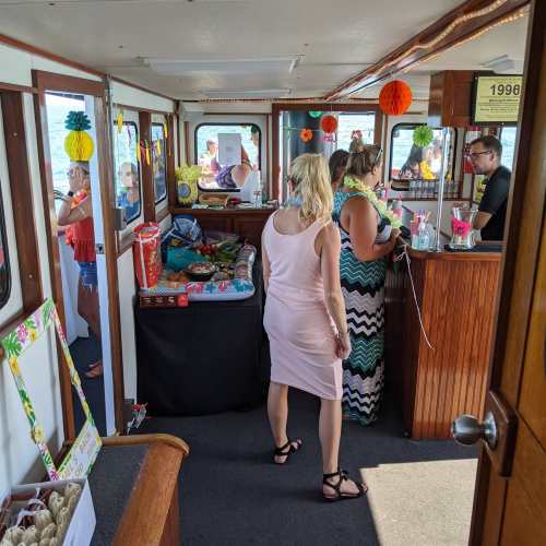 Interior of a boat with people socializing, colorful decorations, and a bar area. Bright and festive atmosphere.