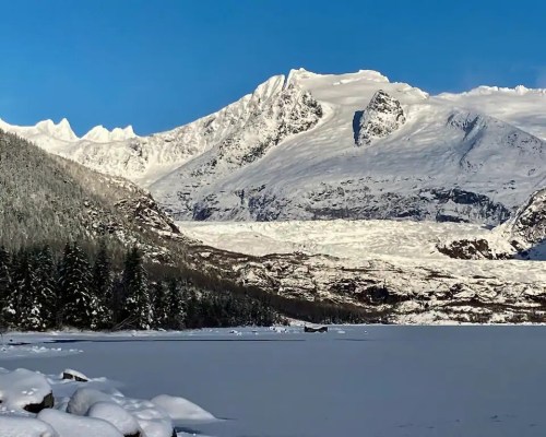 Snow-covered mountains rise above a frozen lake, surrounded by evergreen trees under a clear blue sky.