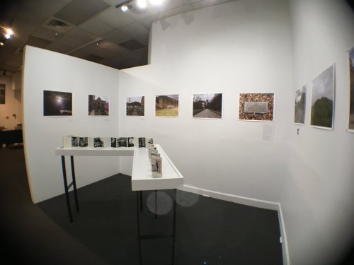 Art gallery interior featuring a white wall with framed photographs and a display table with small books.