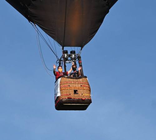 Two people wave from a hot air balloon basket against a clear blue sky.