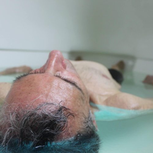 A person with short hair is lying back in a shallow pool of water, appearing relaxed and serene.