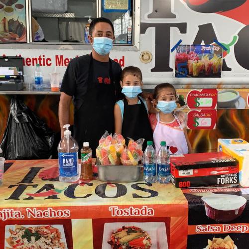 A taco vendor stands with two children at a food stall, all wearing masks, surrounded by colorful snacks and drinks.