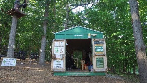 A green building in a forest, labeled "Harnessing," with a "Now Hiring" sign and outdoor activity equipment visible.