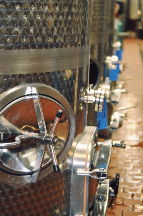 Close-up of stainless steel fermentation tanks with valves and blue tags in a winery setting.