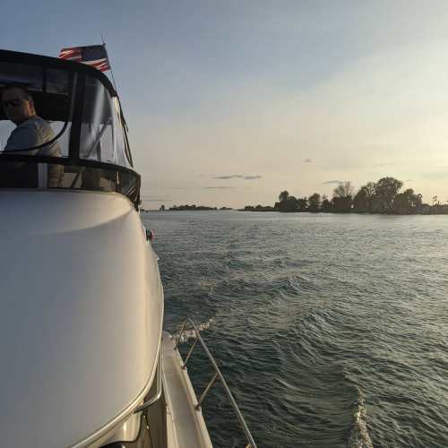 A boat glides through calm waters at sunset, with a person at the helm and trees lining the shore in the distance.