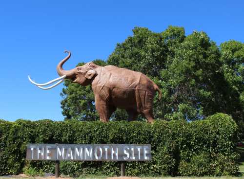 A large mammoth statue stands atop a hedge, with a blue sky and trees in the background, labeled "The Mammoth Site."