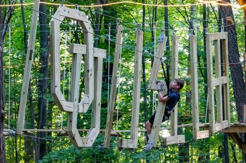 A child climbs on a wooden structure with large letters spelling "OUTSIDE" in a forested area.