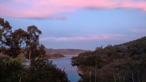 A serene landscape featuring a lake surrounded by hills and trees, with a pastel pink and blue sky at sunset.