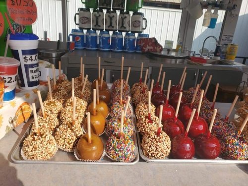 A display of caramel and candy apples, topped with nuts and sprinkles, at a food stand. Soft drinks in the background.
