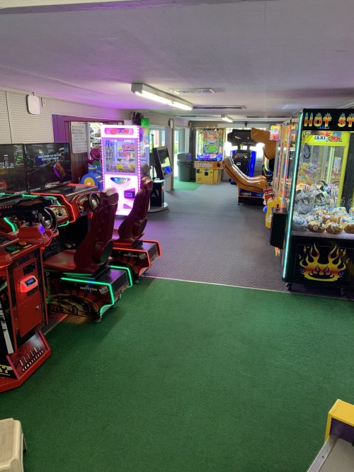 A colorful arcade with various gaming machines, including racing games and claw machines, under bright lights.