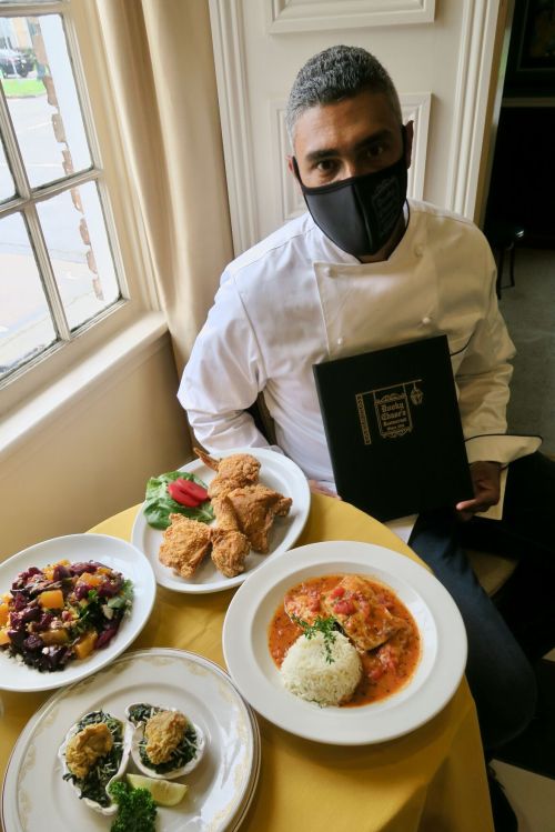 A chef in a mask sits at a table with a menu, surrounded by plates of fried chicken, salad, and stuffed mushrooms.