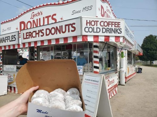 A hand holds a box of donuts in front of Lerch's Donuts stand, featuring a sign for hot coffee and cold drinks.