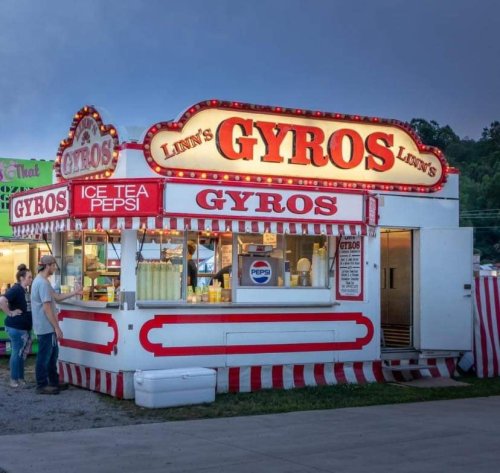 A colorful food stand named "Lynn's Gyros" with bright lights, serving gyros and drinks at a fairground.