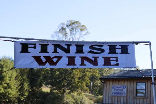 Banner reading "FINISH WINE" displayed at an outdoor event, surrounded by trees and a rustic building.