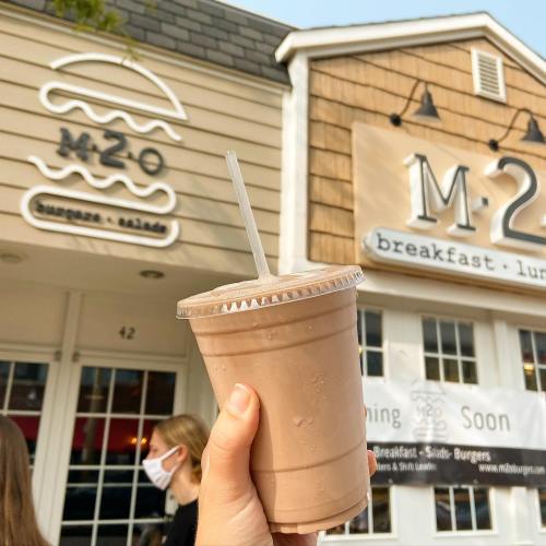 A hand holds a smoothie cup outside a restaurant with signs for breakfast and lunch.