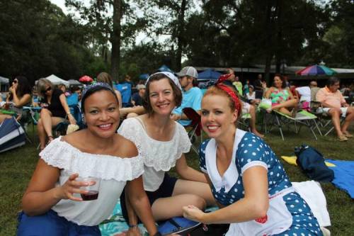 Three women in vintage outfits smile while sitting on the grass at an outdoor event, surrounded by people and tents.