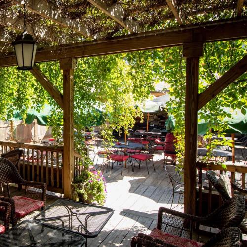 A cozy outdoor dining area with wooden tables, chairs, and lush greenery overhead, creating a shaded atmosphere.