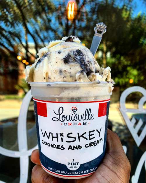 A hand holding a pint of Louisville Cream whiskey cookies and cream ice cream, with a spoon resting on top.
