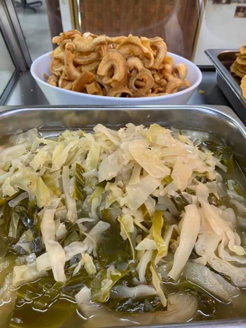 A metal tray with pickled cabbage and greens, alongside a bowl of crispy fried snacks in the background.