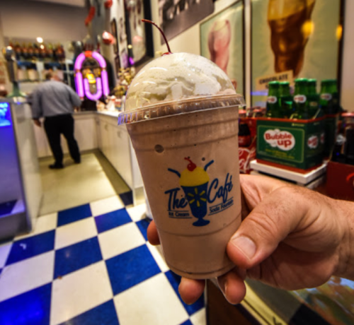 A hand holds a chocolate milkshake topped with whipped cream and a cherry, with a retro café background.