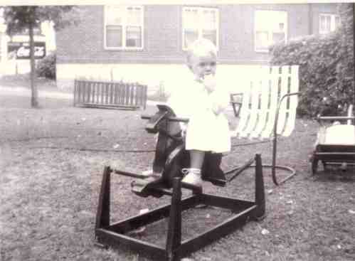 A black and white photo of a child sitting on a rocking horse in a yard, holding something in their mouth.