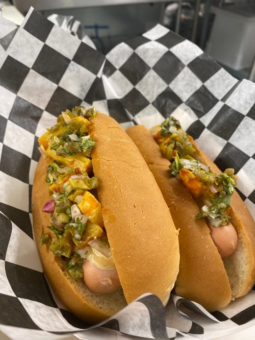 Two hot dogs in a bun topped with chopped vegetables and sauces, served in a black and white checkered paper.