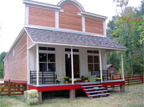 A charming two-story house with a wooden exterior, front porch, and flower pots, surrounded by trees and a wooden fence.