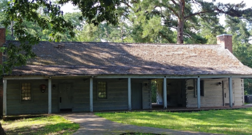 A rustic wooden cabin with a sloped roof, surrounded by trees and a grassy area.