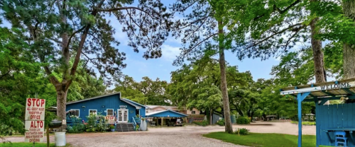 A blue building surrounded by trees, with a gravel path and a sign for an office in a rural setting.