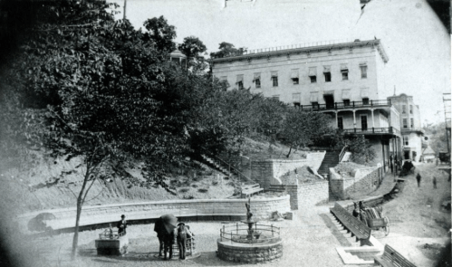 Historic black-and-white photo of a park with steps, benches, and a building in the background. People are gathered.